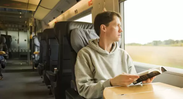 Junger Mann sitzt im Zug, hält ein Buch in der Hand, schaut aus dem Fenster