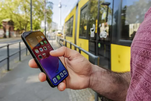 Hand eines Mannes, die Smartphone hält, am Bahngleis stehend, gelbe Bahn fährt ein 